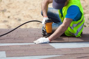 Roofer builder worker with nailgun installing Asphalt Shingles or Bitumen Tiles on a new house under construction