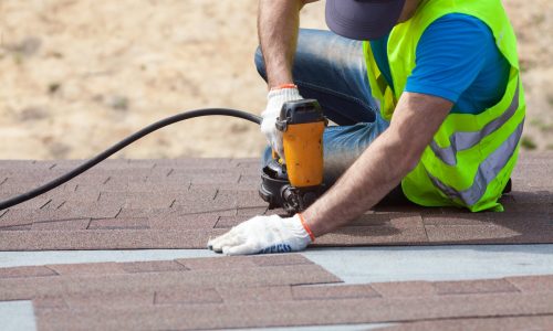 Roofer builder worker with nailgun installing Asphalt Shingles or Bitumen Tiles on a new house under construction